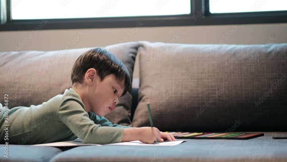Little asian boy having fun on sofa, boy was coloring cartoons on coloring book at home. Coloring Book Education Talent Concept. Copy space. Little asian boy having fun on sofa, boy was coloring cartoons on coloring book at home. Coloring Book Education Talent Concept. Copy space.