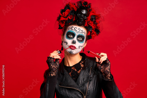 Girl with sugar skull makeup with a wreath of flowers on her head and skull, wearth lace gloves and leather jacket, testing new lip gloss isolated on red background