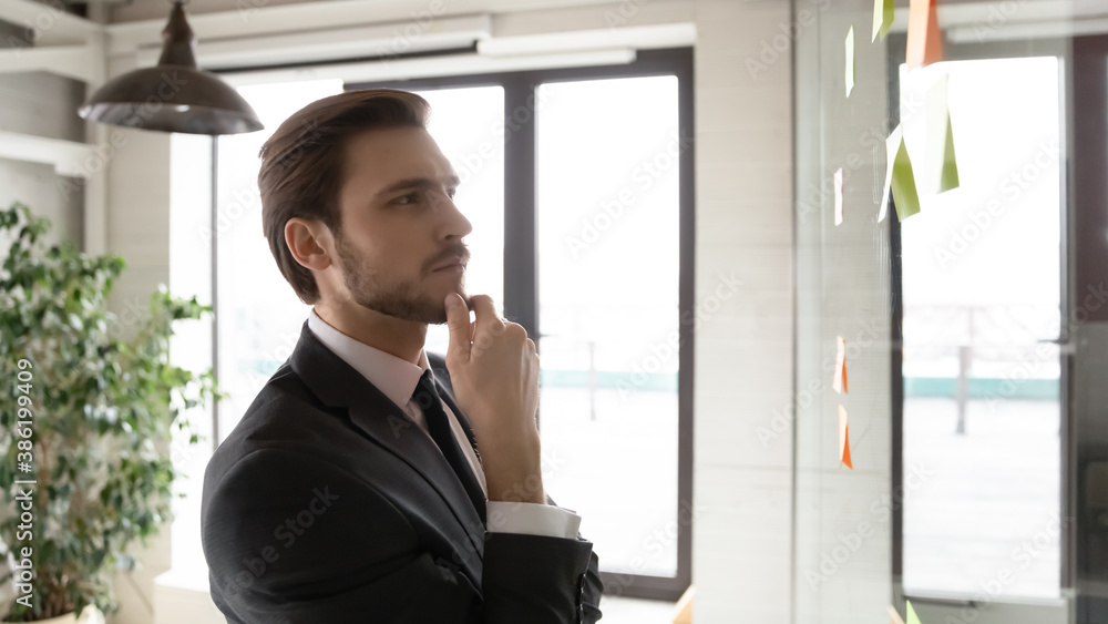 Head shot pensive young corporate employee using project management tool, analyzing steps or checking to do list on sticky paper notes attached to glass wall in modern office, work efficiency concept. Head shot pensive young corporate employee using project management tool, analyzing steps or checking to do list on sticky paper notes attached to glass wall in modern office, work efficiency concept.