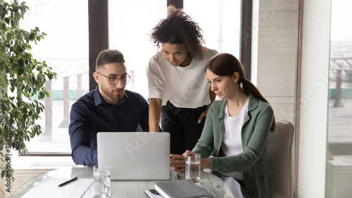 Young arab male manager discussing online project on computer with multiracial female colleagues, brainstorming together at office meeting. Mixed race business people busy with problem solution. photo