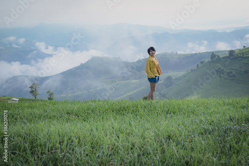 Asian woman was walking comfortably on the top of the hill there is a green grass, a big tree and a cloud on the top of the hill.