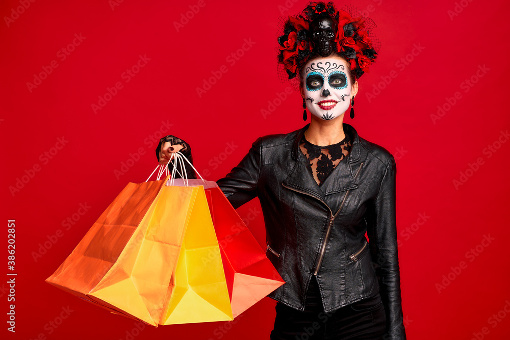 Girl with sugar skull makeup with a wreath of flowers on her head and skull, wearth lace gloves and leather jacket, hold shopper bags party preparation isolated on red background