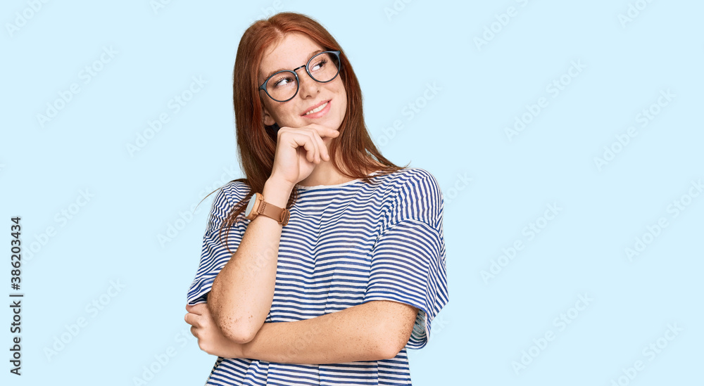Young read head woman wearing casual clothes and glasses with hand on chin thinking about question, pensive expression. smiling and thoughtful face. doubt concept. Young read head woman wearing casual clothes and glasses with hand on chin thinking about question, pensive expression. smiling and thoughtful face. doubt concept.
