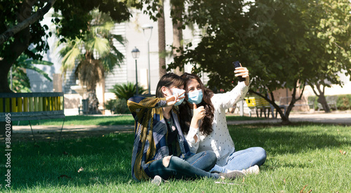  Friends smiling behind the face mask, take a selfie with the smartphone while making the symbol of victory with their hands. Happy friends have a good time outdoors during the new normal.