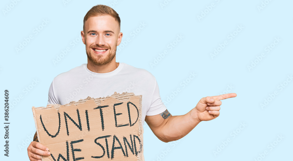 Young irish man holding united we stand banner smiling happy pointing with hand and finger to the side Young irish man holding united we stand banner smiling happy pointing with hand and finger to the side