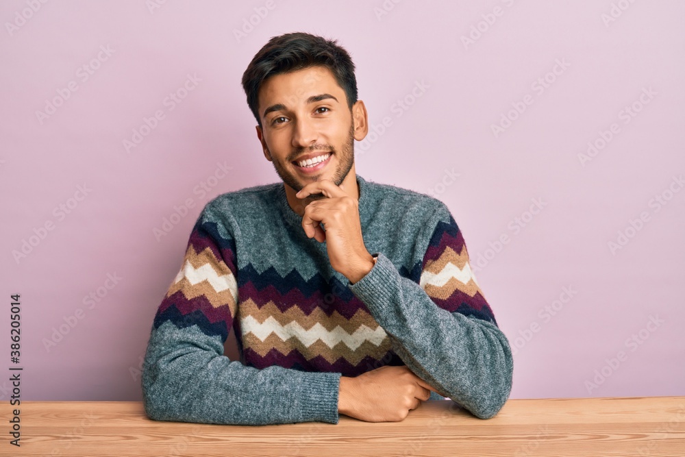 Young handsome man wearing casual winter sweater sitting on the table smiling looking confident at the camera with crossed arms and hand on chin. thinking positive. Young handsome man wearing casual winter sweater sitting on the table smiling looking confident at the camera with crossed arms and hand on chin. thinking positive.