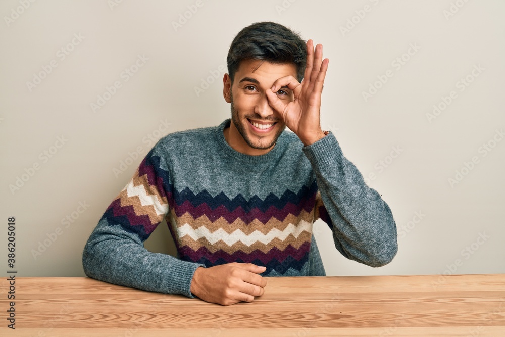 Young handsome man wearing casual sweater sitting on the table doing ok gesture with hand smiling, eye looking through fingers with happy face. Young handsome man wearing casual sweater sitting on the table doing ok gesture with hand smiling, eye looking through fingers with happy face.