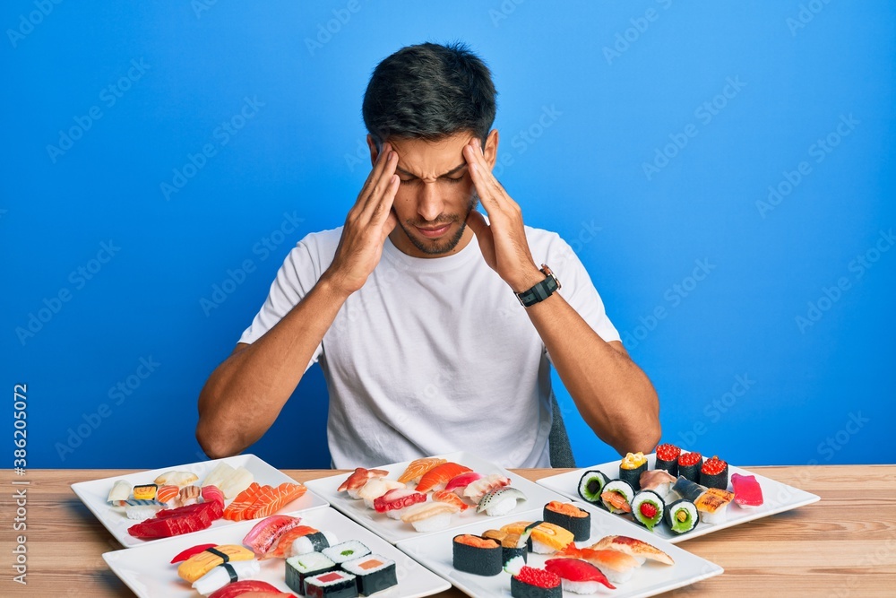 Young handsome man eating sushi sitting on the table with hand on head for pain in head because stress. suffering migraine.