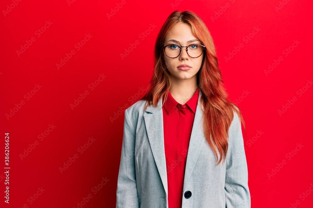Young redhead woman wearing business jacket and glasses with serious expression on face. simple and natural looking at the camera.