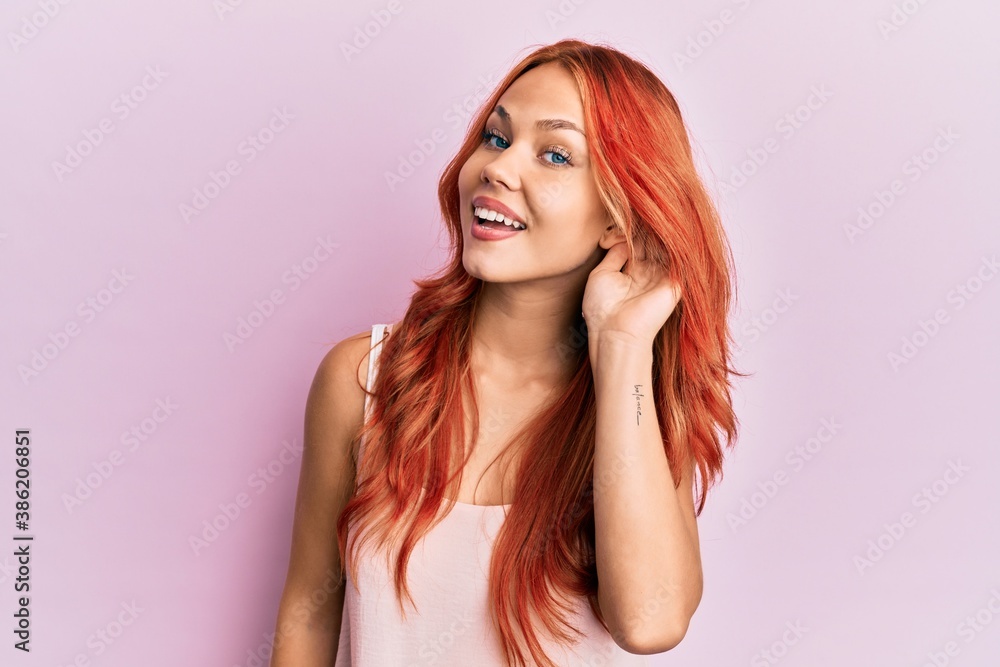 Young beautiful redhead woman wearing casual clothes over pink background smiling with hand over ear listening and hearing to rumor or gossip. deafness concept. Young beautiful redhead woman wearing casual clothes over pink background smiling with hand over ear listening and hearing to rumor or gossip. deafness concept.