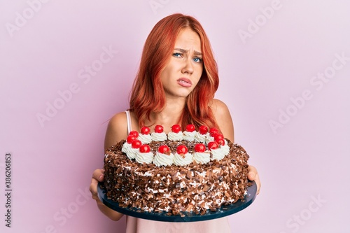 Young redhead woman celebrating birthday with cake clueless and confused expression. doubt concept.