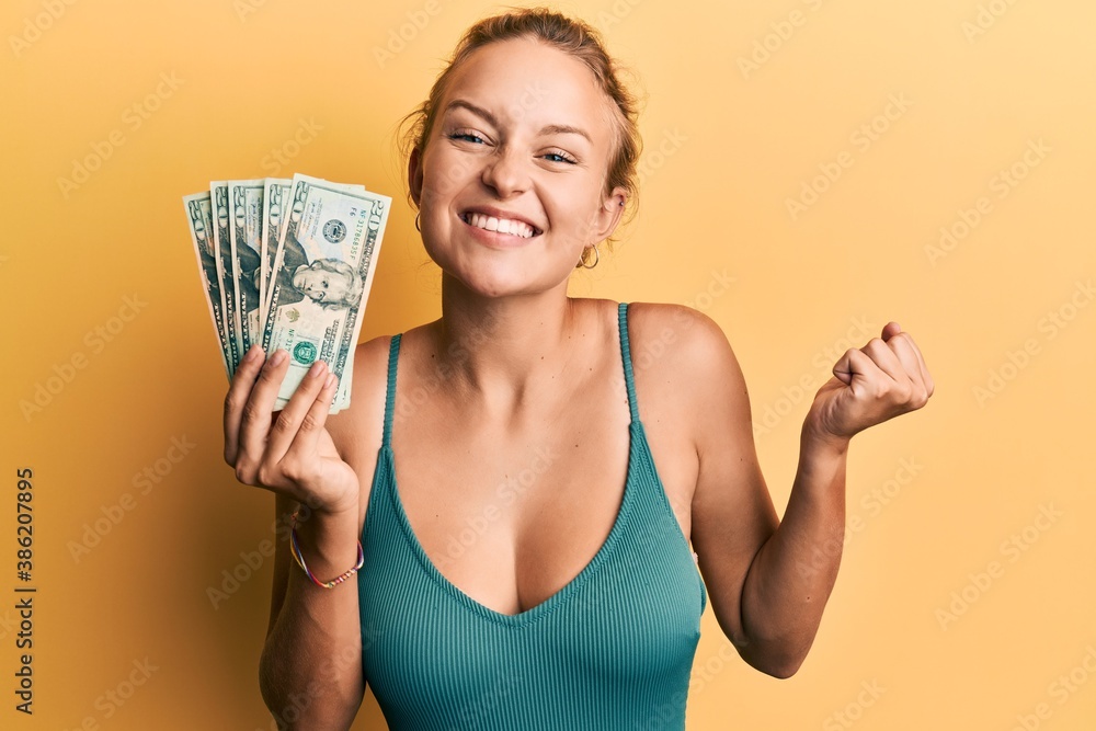 Beautiful caucasian woman holding dollars screaming proud, celebrating victory and success very excited with raised arm Beautiful caucasian woman holding dollars screaming proud, celebrating victory and success very excited with raised arm