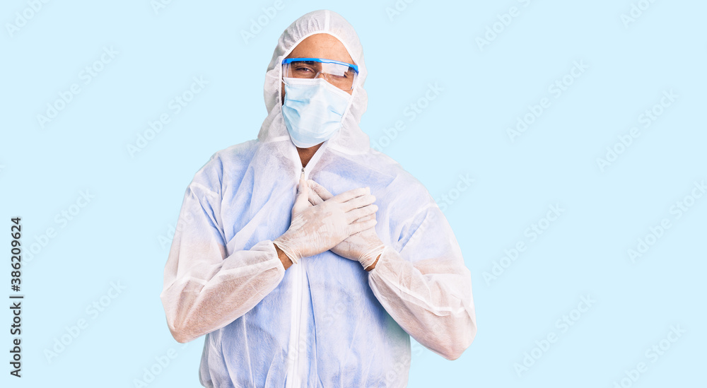 Young hispanic man wearing doctor protection coronavirus uniform and medical mask smiling with hands on chest with closed eyes and grateful gesture on face. health concept. Young hispanic man wearing doctor protection coronavirus uniform and medical mask smiling with hands on chest with closed eyes and grateful gesture on face. health concept.