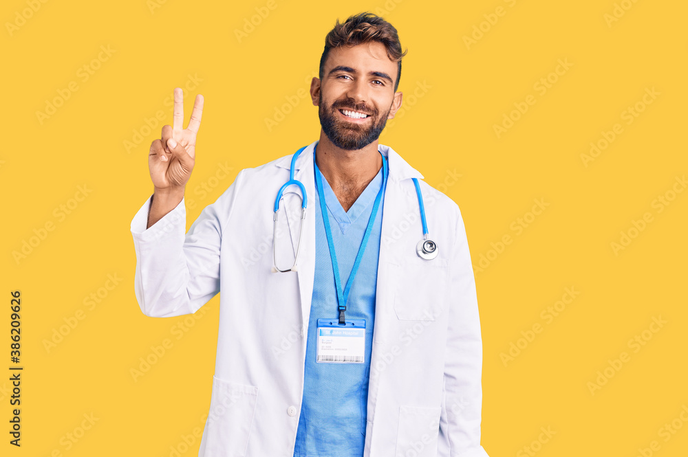 Young hispanic man wearing doctor uniform and stethoscope showing and pointing up with fingers number two while smiling confident and happy.