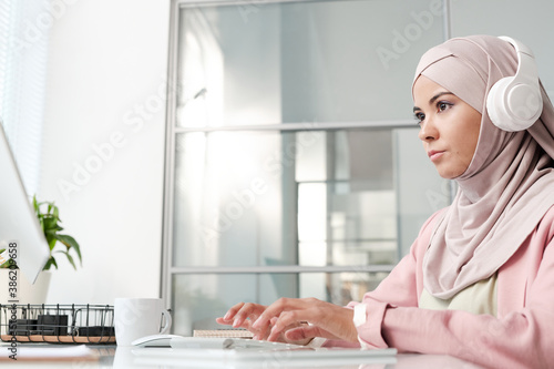 Young serious businesswoman in headphones and hijab looking at computer screen