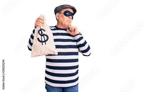 Senior handsome man wearing burglar mask holding money bag serious face thinking about question with hand on chin, thoughtful about confusing idea