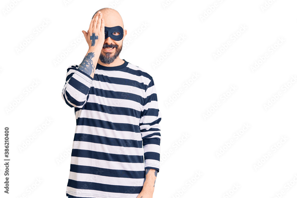 Naklejka premium Young handsome man wearing burglar mask covering one eye with hand, confident smile on face and surprise emotion.