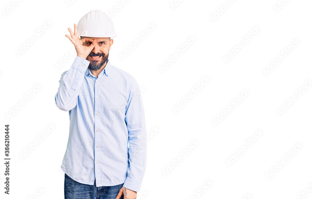 Young handsome man wearing architect hardhat doing ok gesture with hand smiling, eye looking through fingers with happy face. Young handsome man wearing architect hardhat doing ok gesture with hand smiling, eye looking through fingers with happy face.