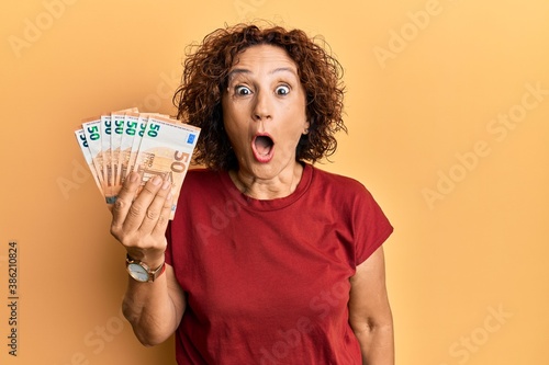 Beautiful middle age mature woman holding bunch of 50 euro banknotes scared and amazed with open mouth for surprise, disbelief face