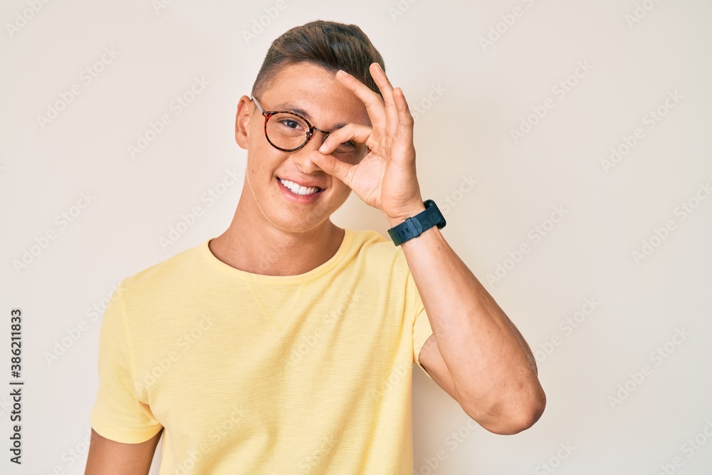 Young hispanic boy wearing casual clothes and glasses smiling happy doing ok sign with hand on eye looking through fingers Young hispanic boy wearing casual clothes and glasses smiling happy doing ok sign with hand on eye looking through fingers