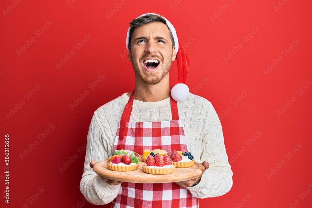 Handsome caucasian man wearing baker apron and christmas hat holding cakes angry and mad screaming frustrated and furious, shouting with anger looking up. Handsome caucasian man wearing baker apron and christmas hat holding cakes angry and mad screaming frustrated and furious, shouting with anger looking up.