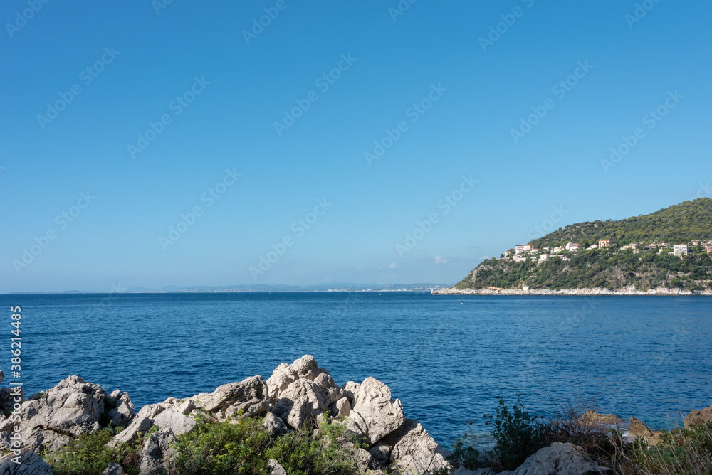 View on the coast of mediterranean sea under blue sky. Rocks in the foreground. Saint-Jean-Cap-Ferrat hiking trail. French Riviera, Cote d'Azur. View on the coast of mediterranean sea under blue sky. Rocks in the foreground. Saint-Jean-Cap-Ferrat hiking trail. French Riviera, Cote d'Azur.
