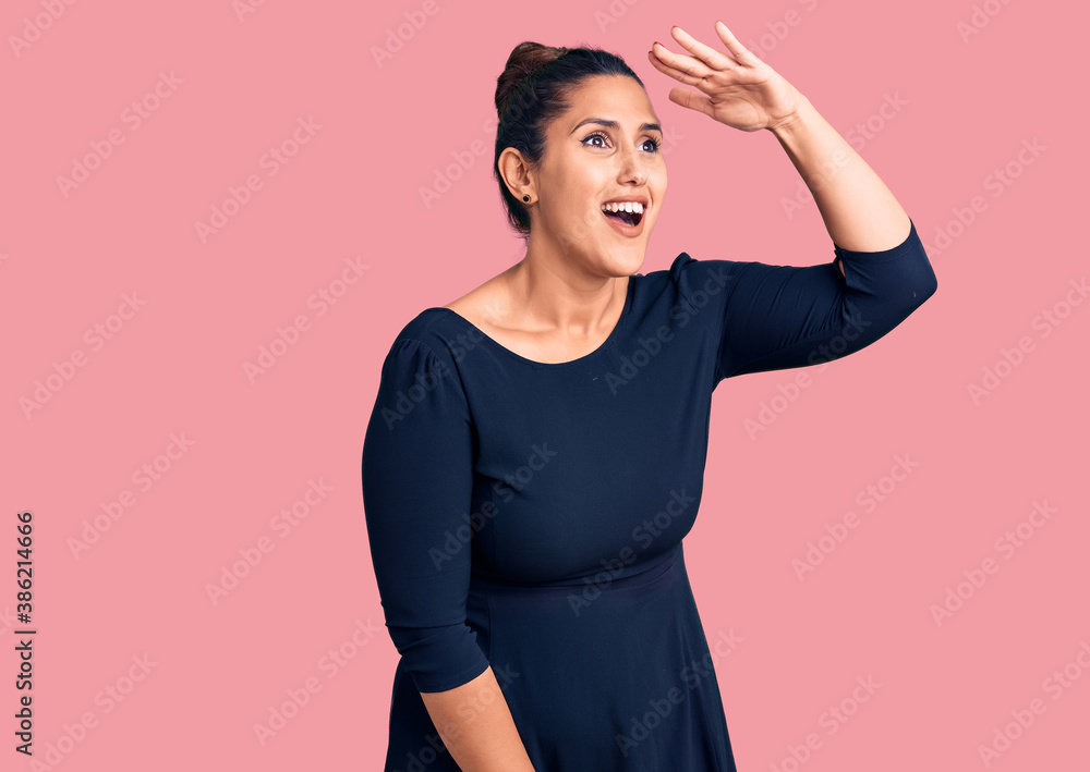 Young beautiful brunette woman wearing casual dress very happy and smiling looking far away with hand over head. searching concept. Young beautiful brunette woman wearing casual dress very happy and smiling looking far away with hand over head. searching concept.
