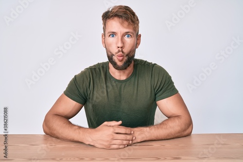 Young caucasian man wearing casual clothes sitting on the table making fish face with lips, crazy and comical gesture. funny expression.