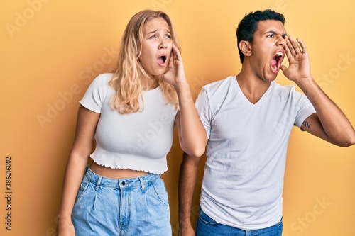 Young interracial couple wearing casual white tshirt shouting and screaming loud to side with hand on mouth. communication concept.