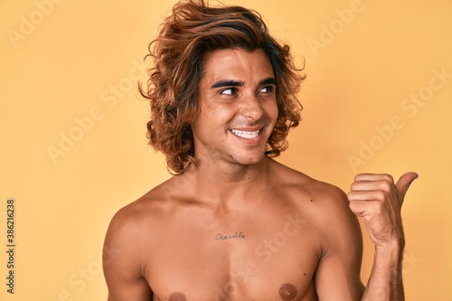 Young hispanic man standing shirtless smiling with happy face looking and pointing to the side with thumb up.