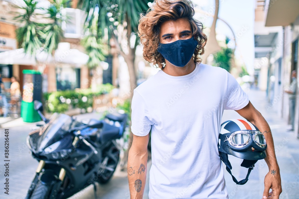 Young hispanic man wearing coronavirus protection mask holding moto helmet over motorcycle at the city