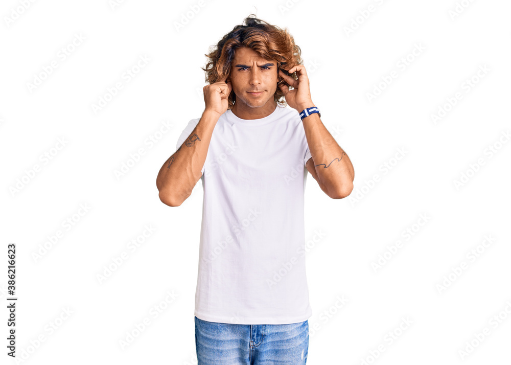 Young hispanic man wearing casual white tshirt covering ears with fingers with annoyed expression for the noise of loud music. deaf concept. Young hispanic man wearing casual white tshirt covering ears with fingers with annoyed expression for the noise of loud music. deaf concept.