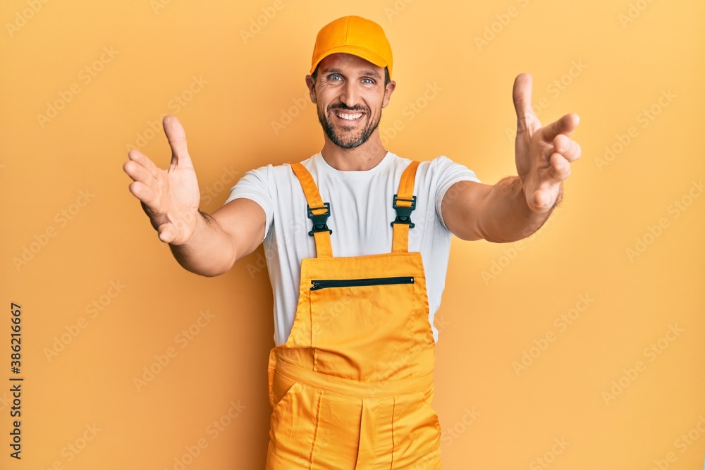 Young handsome man wearing handyman uniform over yellow background looking at the camera smiling with open arms for hug. cheerful expression embracing happiness.