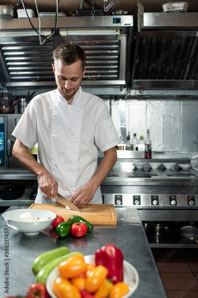 Young professional chef in uniform cutting fresh onion and other vegetables