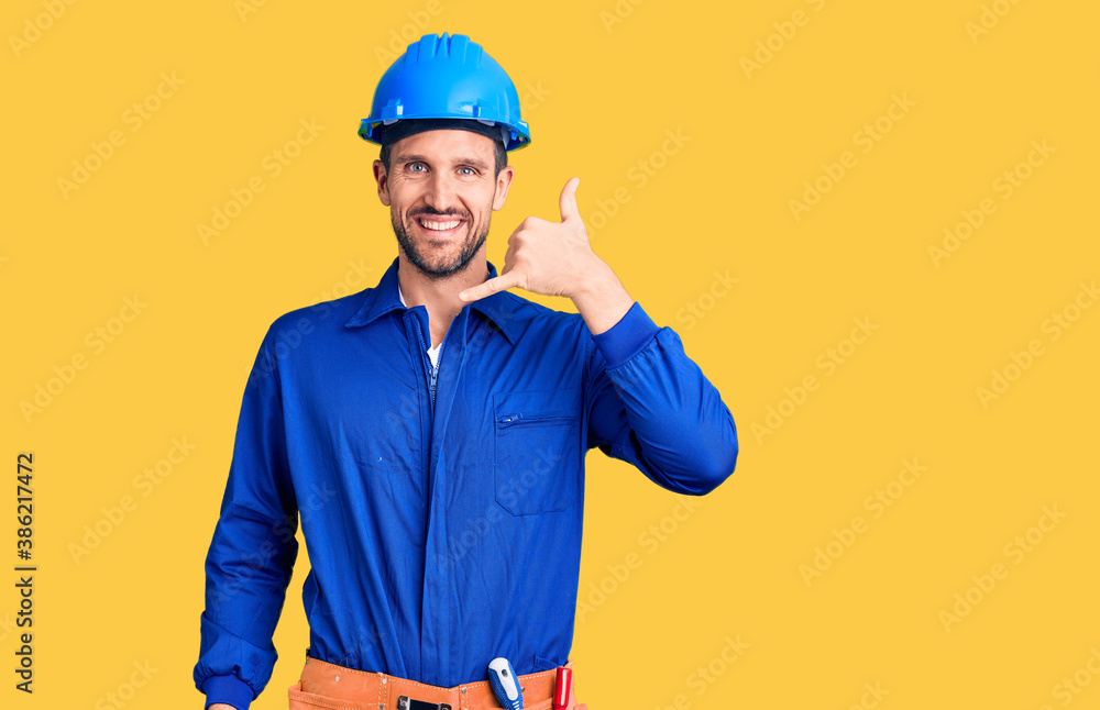 Young handsome man wearing worker uniform and hardhat smiling doing phone gesture with hand and fingers like talking on the telephone. communicating concepts.