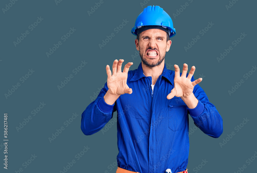 Young handsome man wearing worker uniform and hardhat smiling funny doing claw gesture as cat, aggressive and sexy expression