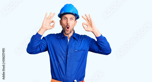 Young handsome man wearing worker uniform and hardhat surprised pointing with hand finger to the side, open mouth amazed expression.