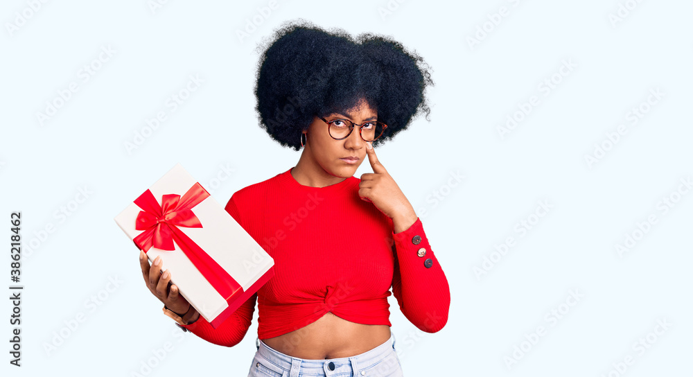 Young african american girl holding gift pointing to the eye watching you gesture, suspicious expression Young african american girl holding gift pointing to the eye watching you gesture, suspicious expression