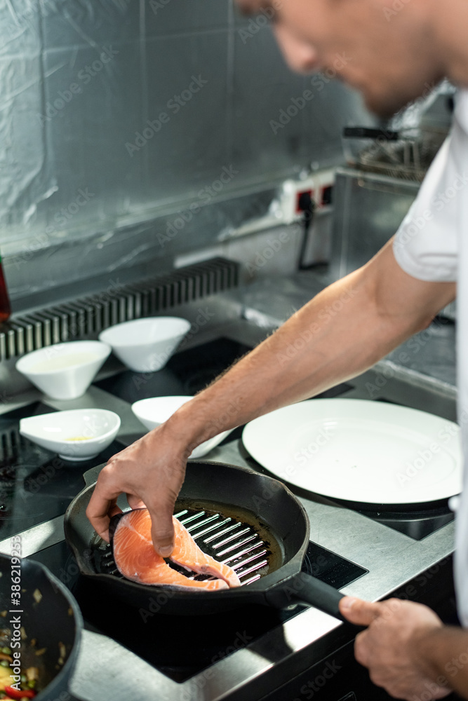 Hand of young chef putting piece of salmon on hot frying pan with olive oil
