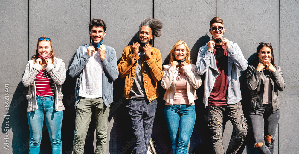 Fototapeta premium Multiracial friends smiling with face mask after lockdown reopening - Happy guys and girls having fun around city streets - University students on sunny day at urban wall - Warm bright filter