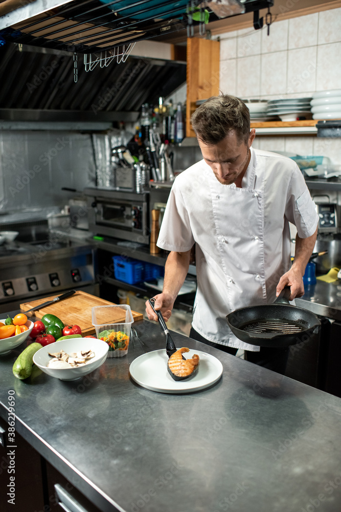Young male chef in white uniform putting piece of fried salmon on plate Young male chef in white uniform putting piece of fried salmon on plate
