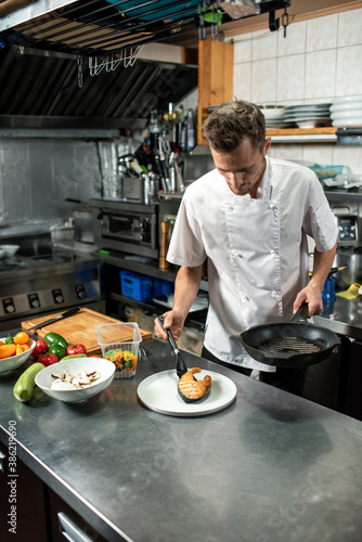 Young male chef in white uniform putting piece of fried salmon on plate