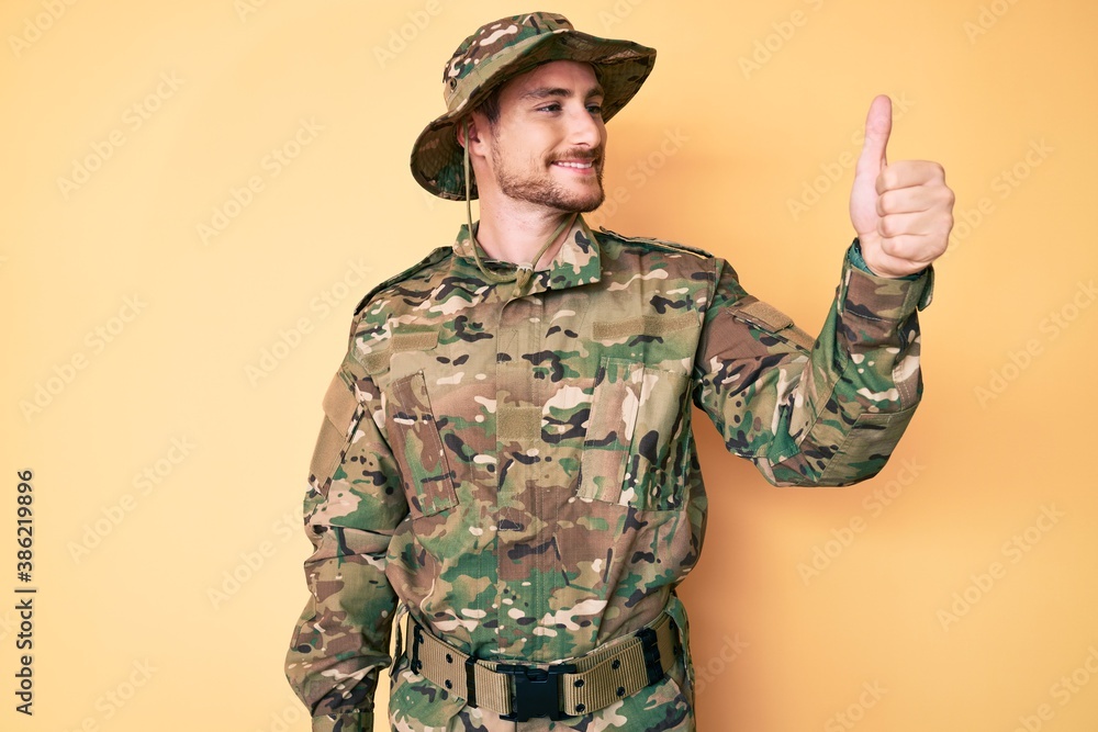 Young caucasian man wearing camouflage army uniform looking proud, smiling doing thumbs up gesture to the side Young caucasian man wearing camouflage army uniform looking proud, smiling doing thumbs up gesture to the side