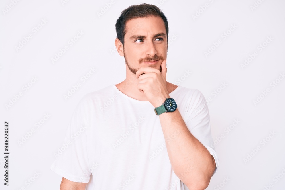 Young handsome man wearing casual white tshirt with hand on chin thinking about question, pensive expression. smiling with thoughtful face. doubt concept. Young handsome man wearing casual white tshirt with hand on chin thinking about question, pensive expression. smiling with thoughtful face. doubt concept.