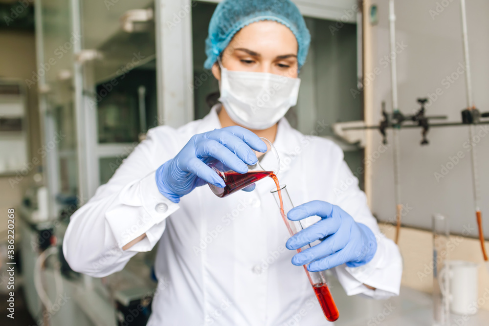 Young female researcher in medical cap and protective mask are doing investigations with test tube red solution in laboratory. Young female researcher in medical cap and protective mask are doing investigations with test tube red solution in laboratory.
