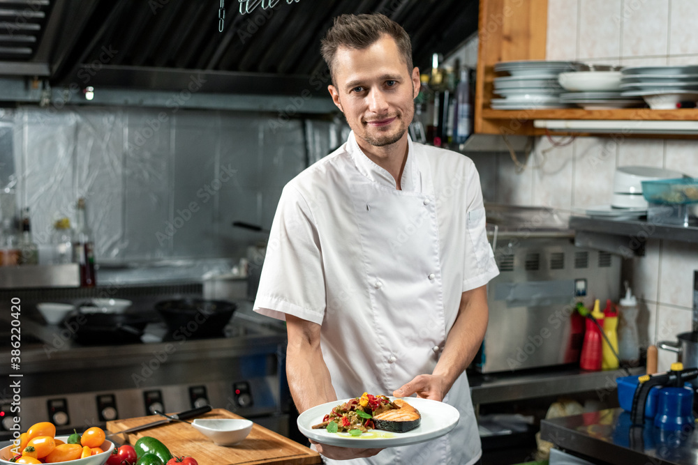 Happy young professional chef in white uniform holding plate with fried salmon Happy young professional chef in white uniform holding plate with fried salmon