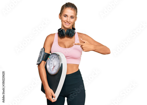 Young blonde woman wearing sportswear holding weighing machine pointing finger to one self smiling happy and proud