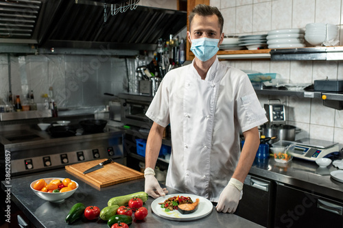 Young professional chef in uniform and protective mask holding fried salmon