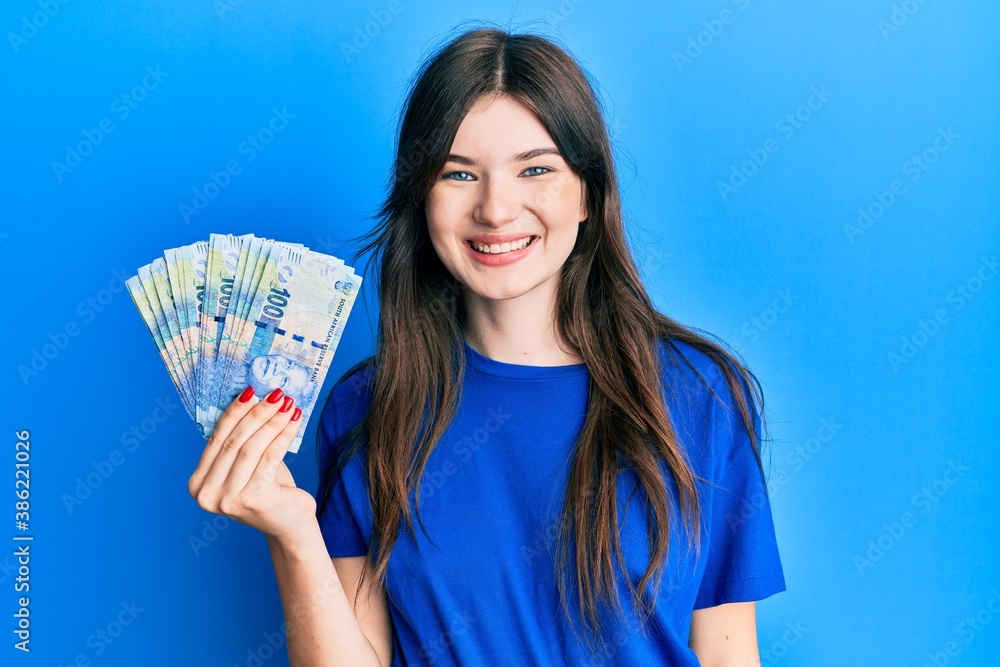 Young beautiful caucasian girl holding south african 100 rand banknotes looking positive and happy standing and smiling with a confident smile showing teeth Young beautiful caucasian girl holding south african 100 rand banknotes looking positive and happy standing and smiling with a confident smile showing teeth