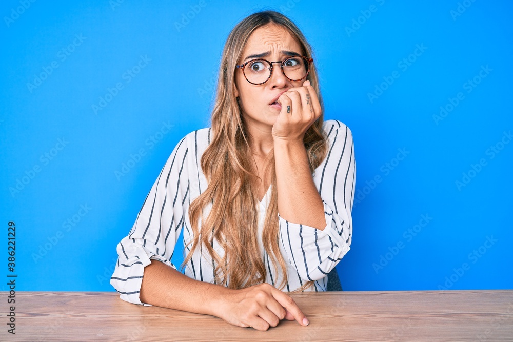Young beautiful blonde woman wearing glasses sitting on the table looking stressed and nervous with hands on mouth biting nails. anxiety problem. Young beautiful blonde woman wearing glasses sitting on the table looking stressed and nervous with hands on mouth biting nails. anxiety problem.